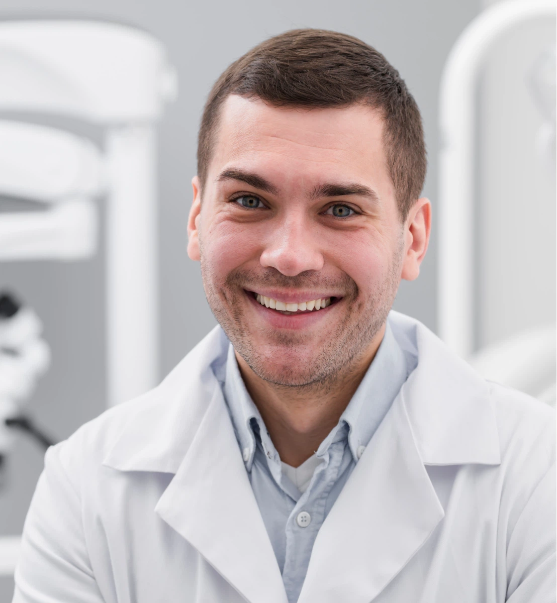 Smiling young male doctor wearing a white coat and light blue shirt in a medical office.